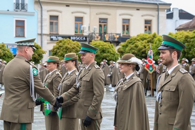 Na zdjęciu widać sześć osób, które ubrane są w mundury. Dwaj mężczyźni podają sobie ręce, na których mają czarne rękawiczki. W tle stoi rząd budynków. Widoczne są zielone drzewa.