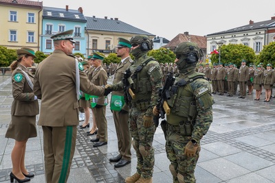 Grupa ludzi ubrana w mundury stoi na płycie rynku. Z przodu stoją osoby ubrane w mundury, dwaj mężczyźni podają sobie ręce, na których mają czarne rękawiczki. Dwóch mężczyzn ma zasłoniętą twarz. Płyty są mokre od deszczu. W tle stoi rząd budynków. Widoczne są zielone drzewa.