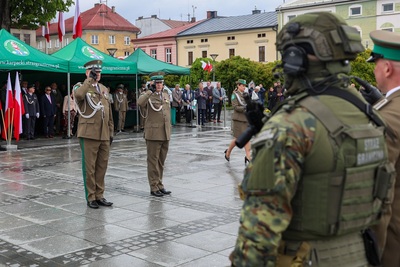 Grupa ludzi ubrana w mundury stoi na płycie rynku. Dwóch mężczyzn oddaje honor pozostałym. Z lewej strony zdjęcia widać cztery zielone namioty pod którymi stoi wiele osób. Na środku widać flagi biało-czerwone. W tle stoi rząd budynków. Widoczne są zielone drzewa. Płyty są mokre od deszczu.