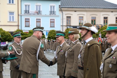 Na zdjęciu widać siedem osób, które ubrane są w mundury. Dwaj mężczyźni podają sobie ręce, na których mają czarne rękawiczki. W tle stoi rząd budynków. Widoczne są zielone drzewa. Z lewej strony jedna osoba w rękach trzyma zieloną podstawkę ma której widać zielone małe pudełka.
