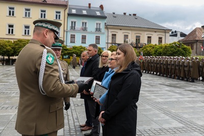 Na zdjęciu dwie osoby w mundurach wręczają tabliczki pięciu osobom. W tle stoi rząd budynków. Widoczne są zielone drzewa. Widać w tle stojącą w rzędzie grupę osób.