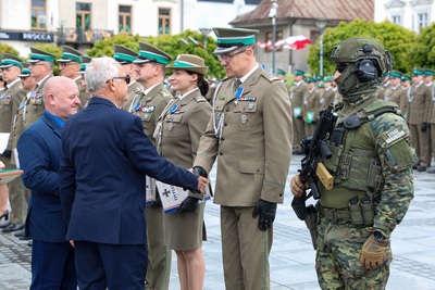 Grupa ludzi ubrana w mundury stoi na płycie rynku. Dwóch mężczyzn ubranych w granatowe marynarki stoi przed nimi. Jeden z nich podaje rękę drugiemu mężczyźnie ubranemu w mundur. W tle stoi rząd budynków. Widoczne są zielone drzewa. Widać w tle stojącą w rzędzie grupę osób.