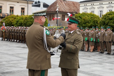 Dwóch mężczyzn w mundurach trzyma w rękach szklany puchar. W tle stoi rząd budynków. Widoczne są zielone drzewa. Widać w tle stojącą w rzędzie grupę osób.