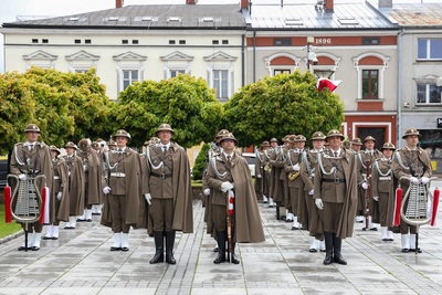 Na zdjęciu widać grupę osób ubraną w mundury i peleryny, na głowach mają kapelusze z piórkiem. W rękach trzymają różne instrumenty. W tle stoi rząd budynków. Widoczne są zielone drzewa na jednym z nich po prawej stronie wetknięta jest flaga biało-czerwona.