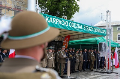 Na pierwszym planie widać zamazaną osobę ubraną w mundur, na głowie ma kapelusz z piórkiem. W tle pod zielonym dachem z napisem Karpacki Oddział Straży Granicznej stoją osoby, jedna z nich przemawia z mównicy. W tle widać budynek i wysokie zielone drzewo. Z prawej strony zdjęcia widać dwie flagi biało-czerwone.