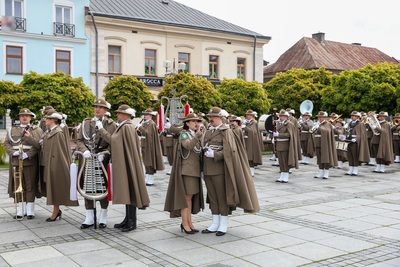 Na zdjęciu widać grupę osób ubraną w mundury i peleryny, na głowach mają kapelusze z piórkiem. W rękach trzymają różne instrumenty. Sześć osób z przodu zdjęcia śpiewa do mikrofonów. W tle stoi rząd budynków. Widoczne są zielone drzewa.