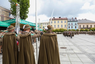 Z przodu zdjęcia widać trzy osoby ubrane w mundury, mają peleryny i kapelusze z piórkiem. Z lewej strony widać dachy zielonych namiotów, na jednym  widać z napis Karpacki Oddział Straży Granicznej. Z prawej strony zdjęcia widać dużą grupę osób w mundurach, która maszeruje. W tle stoi rząd budynków. Widoczne są zielone drzewa.