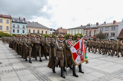 Grupa osób ubrana w mundury maszeruje po płycie rynku. Jedna osoba z przodu trzyma w rękach sztandar w kolorach biało-czerwonych. W tle stoi rząd budynków. Widoczne są zielone drzewa.