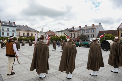 Na zdjęciu widać grupę osób ubraną w mundury, w rękach trzymają  instrumenty. Z lewej strony zdjęcia widać osobą ubraną w strój góralski. W tle widać budynki w szeregu i zielone drzewa.