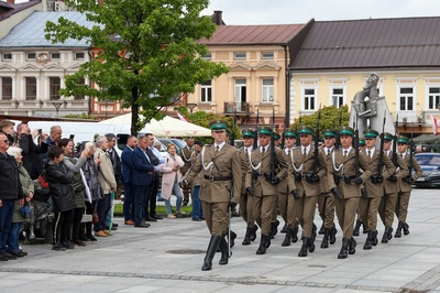 Grupa osób w mundurach maszeruje, z lewej strony widać osoby, które to obserwują. Za nimi widać popielaty pomnik, zielone drzewo i rząd budynków.