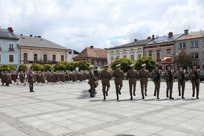 Na zdjęciu widać pokaz musztry paradnej wykonywanej przez mężczyzn w mundurach.  Z prawej strony widać osoby w mundurach z instrumentami w rękach.  W tle widać budynki w szeregu i zielone drzewa.