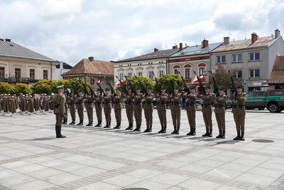 Na zdjęciu widać grupę mężczyzn ubranych w mundury, w rękach trzymają karabinki. W tle widać budynki w szeregu i zielone drzewa oraz samochód koloru zielonego z napisem Straż Graniczna.