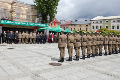 Na zdjęciu widać grupę trzynastu mężczyzn ubranych w mundury, którzy stoją frontem do grupy osób, która znajduje się pod zielonym dachem z napisem Karpacki Oddział Straży Granicznej. W tle widać budynki w szeregu i zielone drzewa.