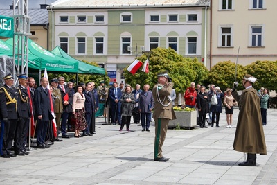 Na zdjęciu widać grupę ludzi, dwóch z nich stoi w mundurach jeden oddaje honor, a drugi w ręku trzyma metalową szablę. W tle widać budynki w szeregu i zielone drzewa.