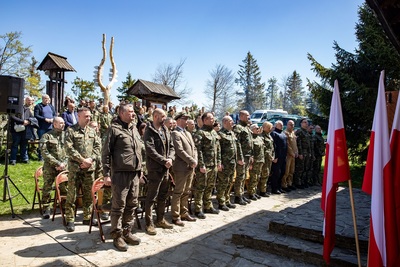 Na zdjęciu widać grupę osób ubranych w mundury, którzy stoją na kamiennej podłodze. Za nimi widać też grupę osób ubranych w zwykłe ciuchy, którzy stoją na trawie. Z prawej strony zdjęcia widać flagi biało-czerwone. W tle widać drzewa iglaste koloru zielonego.