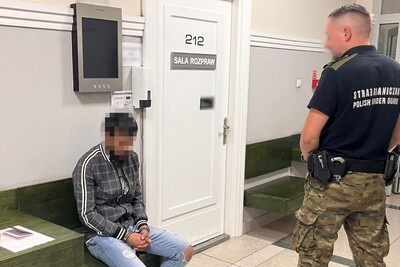 Zatrzymany mołdawianin siedzi na krześle przed salą rozpraw tarnowskiego sądu rejonowego. pilnuje go funkcjonariusz straży granicznej. cudzoziemiec ma skute kajdankami ręce