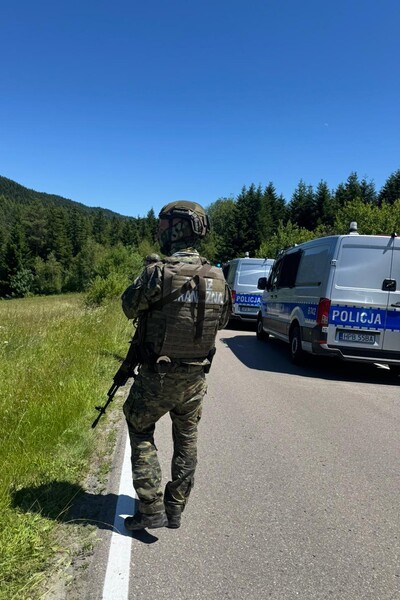 Funkcjonariusz straży granicznej ubrani w kamizelki kuloodporne i hełmy idący obok samochodów służbowych policji