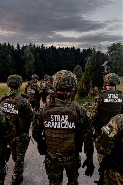 Funkcjonariusze straży granicznej ubrani w kamizelki kuloodporne i hełmy zmierzający w stronę lasu o zachodzenie słońca