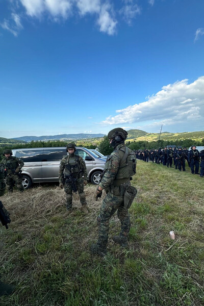 Funkcjonariusz straży granicznej ubrani w kamizelki kuloodporne i hełmy rozmawiający ze sobą w tle widok na góry oraz duża ilość policjantów idąca w ich stronę