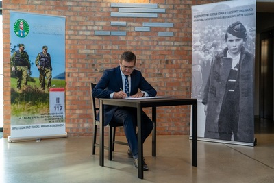 Zastępca Dyrektora  muzeum Auschwitz podczas podpisywania porozumienia siedzi na krześle przy stole ubrany w garnitur  w tle rollapay karpackiego oddziału straży granicznej i muzeum Auschwitz