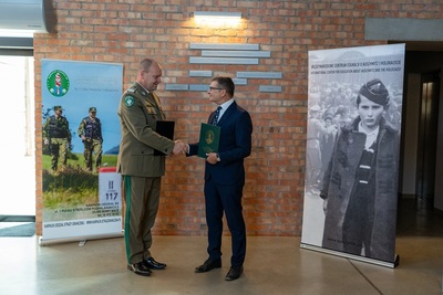Komendant Karpackiego Oddziału straży granicznej i zastępca Dyrektora  muzeum Auschwitz podają sobie dłonie po podpisaniu porozumienia mężczyźni stoją na tle rollapay karpackiego oddziału straży granicznej i muzeum Auschwitz