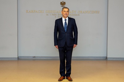 Na zdjęciu widać mężczyznę ubranego w czarny garnitur, stoi na tle białej ściany na której widać napisy złotymi literami