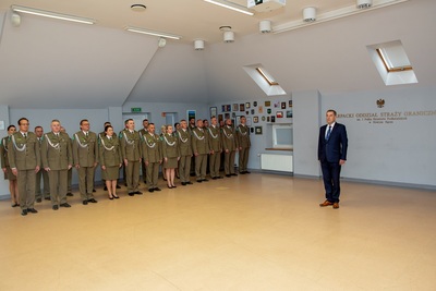 Na zdjęciu po lewej stronie widać grupę osób ubranych w mundury galowe, po prawej stronie stoi mężczyzna w garniturze.