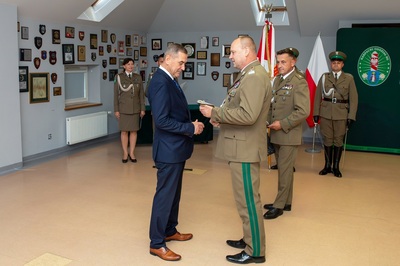 Na zdjęciu widać na pierwszym planie dwóch mężczyzn, jeden ubrany w garnitur, drugi w mundur i wręcza mu ciupagę. W tle widać ludzi w mundurach.