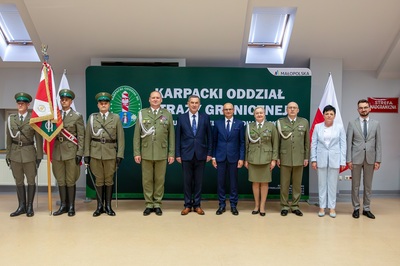 Grupa osób stoi na środku sali, Część z nich ubrana jest w mundury, kilka osób jest w ubraniach cywilnych.  Za nimi znajduje si e duży zielony baner z napisem białymi literami.