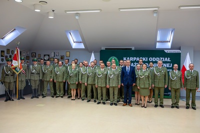 Grupa osób ubrana w mundury galowe stoi na środku pomieszczenia. Wśród nich w samym centrum stoi mężczyzna w garniturze.
