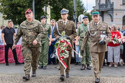Na zdjęciu widać trzech mężczyzn w mundurach. Mężczyzna w środku niesie wieniec, który złożą pod pomnikiem, mężczyzna z p[prawej niesie znicz.