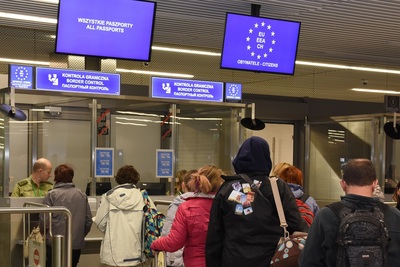 Na zdjęciu widać dużą halę na której u góry wiszą dwie tablice informacyjne z napisem odprawa paszportowa w języku polskim i angielskim. Za szklaną szybą siedzi funkcjonariusz Straży Granicznej w mundurze dokonujący odprawy paszportowej. Przed okienkiem widać wielu podróżnych którzy czekają na odprawę.