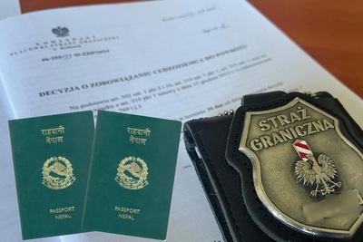 Na zdjęciu widać decyzję o zobowiązaniu cudzoziemca do powrotu, na dokumencie leżą po lewej stronie dwa zielone paszporty, po prawej stronie widać czarną skórzaną legitymację funkcjonariusza z metalowym emblematem na którym widnieje napis straż graniczna biało czerwony słup graniczny i orzeł.
