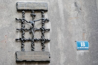 Tablica pamiątkowa Gestapo
Na ścianie budynku znajduje się żeliwna tablica w kształcie kraty więziennej. Pośrodku kraty widoczna jest stylizowana postać ukrzyżowanego człowieka.
Nad kratą znajduje się napis:
„W latach 1940–1945 w kamienicach nr 18 i 20 mieściła się siedziba niemieckiej policji politycznej Gestapo.”
Pod kratą drugi napis:
„Pamięci tych domów, związanych na zawsze z niemieckimi śledztwami więzienia polskich patriotów, z których wielu zamordowano podczas okrutnego śledztwa. Zginęli, abyśmy mogli żyć wolni.”
Obok na ścianie niebieska tabliczka adresowa z białym napisem: „18 ul. Urszulańska”.
