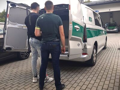 Na zdjęciu samochód Straży Granicznej w kolorze biało - zielonym z otwartymi drzwiami stojący na placu. Obok pojazdu stoi dwóch mężczyzn - funkcjonariusz w zielonej koszulce i dżinsach, obok niego drugi mężczyzna w jasnych spodniach i ciemnej bluzie, którego funkcjonariusz wprowadza do środka radiowozu. W tle widać budynek garażowy a w nim zaparkowany oznakowany samochód służbowy.