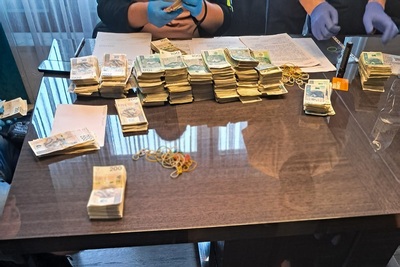 na stole leżą pliki banknotów polskich w różnych nominałach, dokumenty, gumki recepturki. za stołem widać tylko ręce funkcjonariuszy w gumowych rękawiczkach, piszących protokół z zatrzymania tych rzeczy