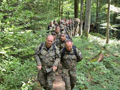 Kilkadziesiąt osób idzie w kolumnie, ale tym razem zdjęcie jest od przodu – widać ich twarze. Pierwsi w kolejce uśmiechają się. Ścieżka jest dość wąska, a po obu stronach rośnie gęsty zielony las.
