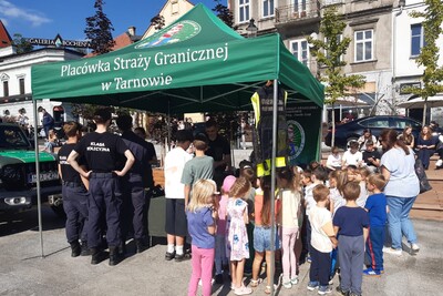 Na zdjęciu zielony namiot wystawowy, z napisem Placówka Straży Granicznej w Tarnowie. Namiot stoi na placu prawdopodobnie w rynku. Po prawej stronie grupa dzieci w kolorowych ubrankach ogląda to, co znajduje się pod namiotem. Po lewej stronie grupa uczniów starszych klas w umundurowaniu charakterystycznym dla klas mundurowych.