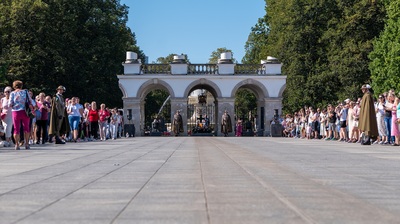 Dwóch żołnierzy w historycznych mundurach w kolorze khaki stoi na baczność po obu stronach Grobu Nieznanego Żołnierza w Warszawie. Mają na sobie peleryny, kapelusze podhalańskie i czarne buty. Każdy z nich trzyma karabin oparty o ziemię. W tle za nimi widać płonący znicz i liczne wieńce w barwach biało-czerwonych. Za żelazną kratą grobu stoją turyści, przyglądający się wartownikom.