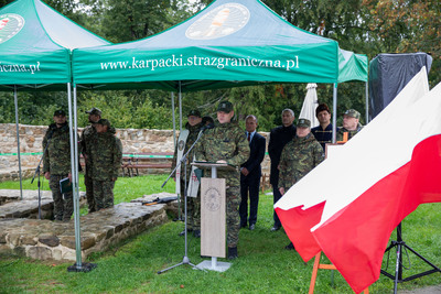 Moment złożenia przysięgi: czworo młodych funkcjonariuszy Straży Granicznej w mundurach, z mieczami w dłoniach, wyciąga prawe ramiona, wskazując palcami do przodu. W tle sztandar i inni żołnierze pod namiotami.