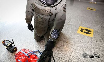 Bliskie ujęcie od pasa w dół na osobę ubraną w ciężki kombinezon ochronny, prawdopodobnie saperski lub do neutralizacji ładunków wybuchowych (EOD), klęczącą na szarej, płytkowanej podłodze. Na przedramieniu kombinezonu widoczny jest zamontowany kontroler z ekranem dotykowym i przyciskami. Obok leży czerwono-czarna torba i małe, elektroniczne urządzenie z anteną.