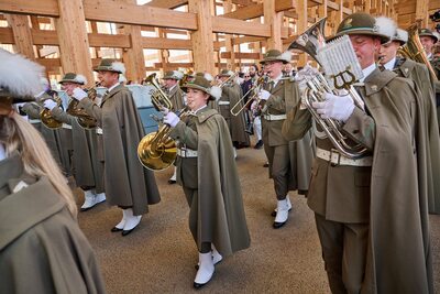 Orkiestra dęta w historycznych, oliwkowych mundurach i pelerynach, z kobietą grającą na waltorni w centrum.