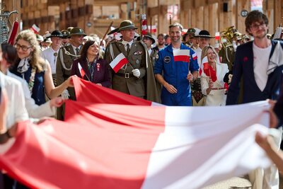 Ludzie trzymający wielką polską flagę, za nimi uśmiechnięty astronauta i delegacja z małymi flagami.