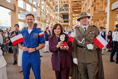 Trzy osoby trzymające polskie flagi: astronauta w niebieskim kombinezonie, kobieta w bordowym kostiumie z maskotką i oficer Straży Granicznej w mundurze.