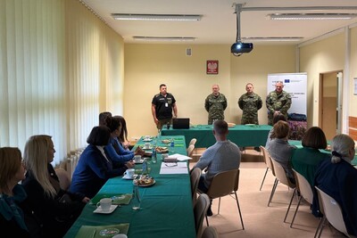 To zdjęcie jest podobne do pierwszego, jednak jest zrobione z nieco innego kąta. Widać tę samą grupę ludzi siedzących przy dwóch stołach. Po prawej stronie wciąż stoją czterej mężczyźni, ale ich pozycja jest lekko zmieniona. W tle za nimi widoczny jest duży, srebrny puchar.