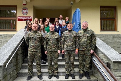 Grupa ludzi pozuje do zdjęcia na schodach przed budynkiem. Na górze schodów, w dwóch rzędach, stoją kobiety i mężczyźni ubrani w odzież cywilną. Na przodzie, na dolnym stopniu, stoi czterech mężczyzn w wojskowych mundurach. W tle widoczna jest fasada budynku z tablicą informacyjną, na której znajduje się herb Polski.