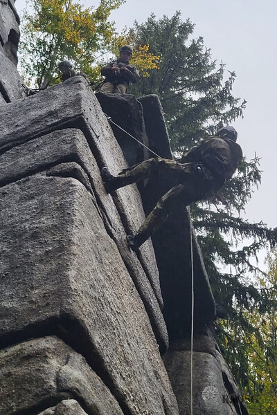 Na zdjęciu widać wysoką skałę po lewej stronie po której wspina się mężczyzna na linie. Na szczycie skały stoją dwie osoby i go asekurują. Z prawej strony zdjęcia widać wysokie drzewa.