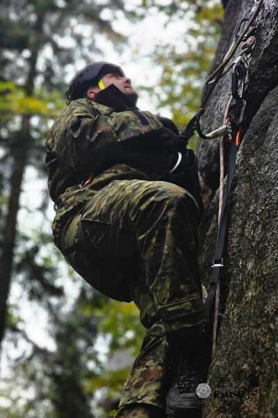 Na zdjęciu widać mężczyznę, ubrany jest w mundur, uprzęż i kask na głowie. Wspina się po wysokiej skale , trzyma przytwierdzoną do skały linę. Pada deszcz, w tle widać z lewej strony drzewa.