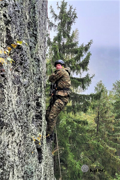 Na zdjęciu widać mężczyznę, ubrany jest w mundur, uprzęż i kask na głowie. Wspina się po wysokiej skale , trzyma przytwierdzoną do skały linę. Z lewej strony zdjęcia widać wysokie zielone drzewa.