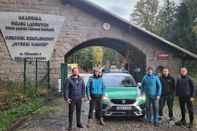 Na zdjęciu widać pięciu mężczyzn ubranych w sportowe ciuchy, po środku stoi samochód koloru srebrnego z zielonymi pasami i napisami. W tle widać bramę wjazdową z wielkim wjazdem w kształcie łuku. Po lewej stronie widać ogrodzenie a na murze tablica informacyjna. Za bramą widać duże podwórze i część zaparkowanej tam karetki w kolorze żółtym i z czerwonymi napisami.